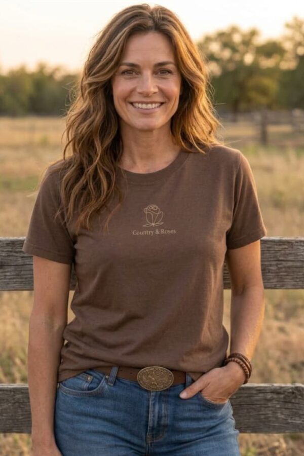 Mujer sonriendo en un rancho luciendo camiseta western marrón de manga corta con logo frontal de rosa